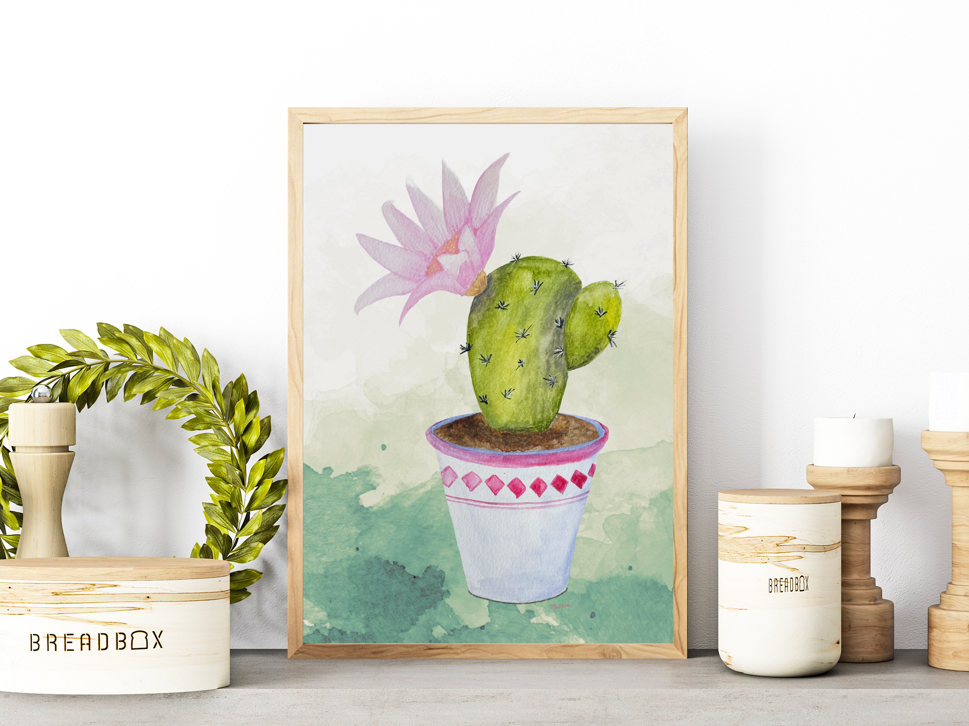 Framed artwork of a potted cactus with a pink flower leaning against a white wall.