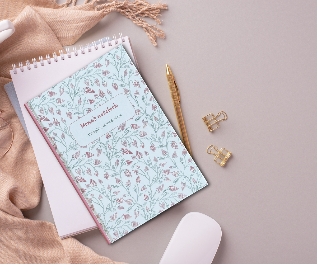 A hardback notebook with a pale green and pink botanical patterned cover labeled 'Mia's notebook' is arranged on a neutral beige surface with a gold pen, white mouse, paper clips, and soft beige fabric.