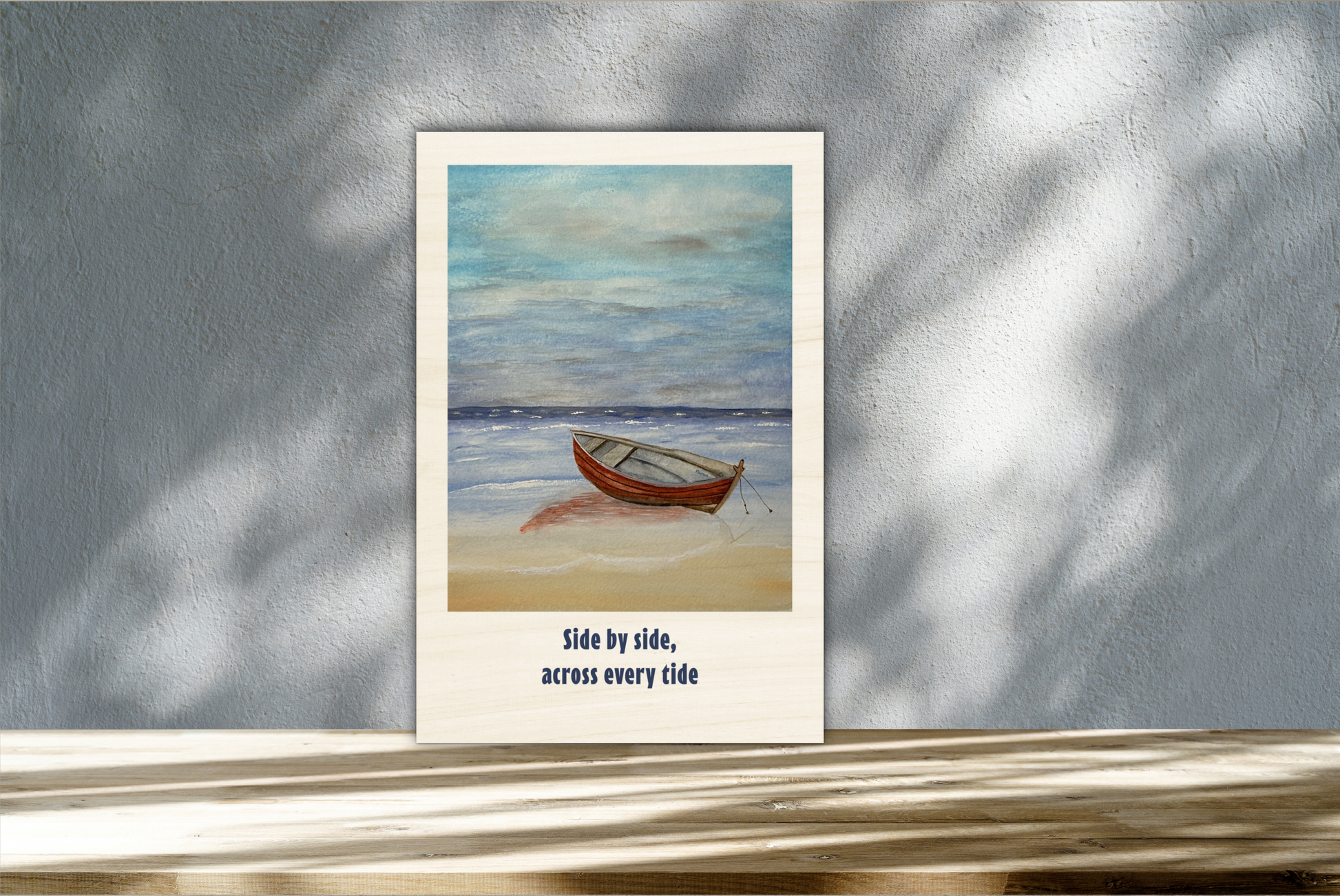 A vertical art print featuring a painting of a small wooden rowboat on a sandy beach under a cloudy blue sky, with the text 'Side by side, across every tide' below. The print is displayed leaning against a textured gray wall with soft leaf shadows.