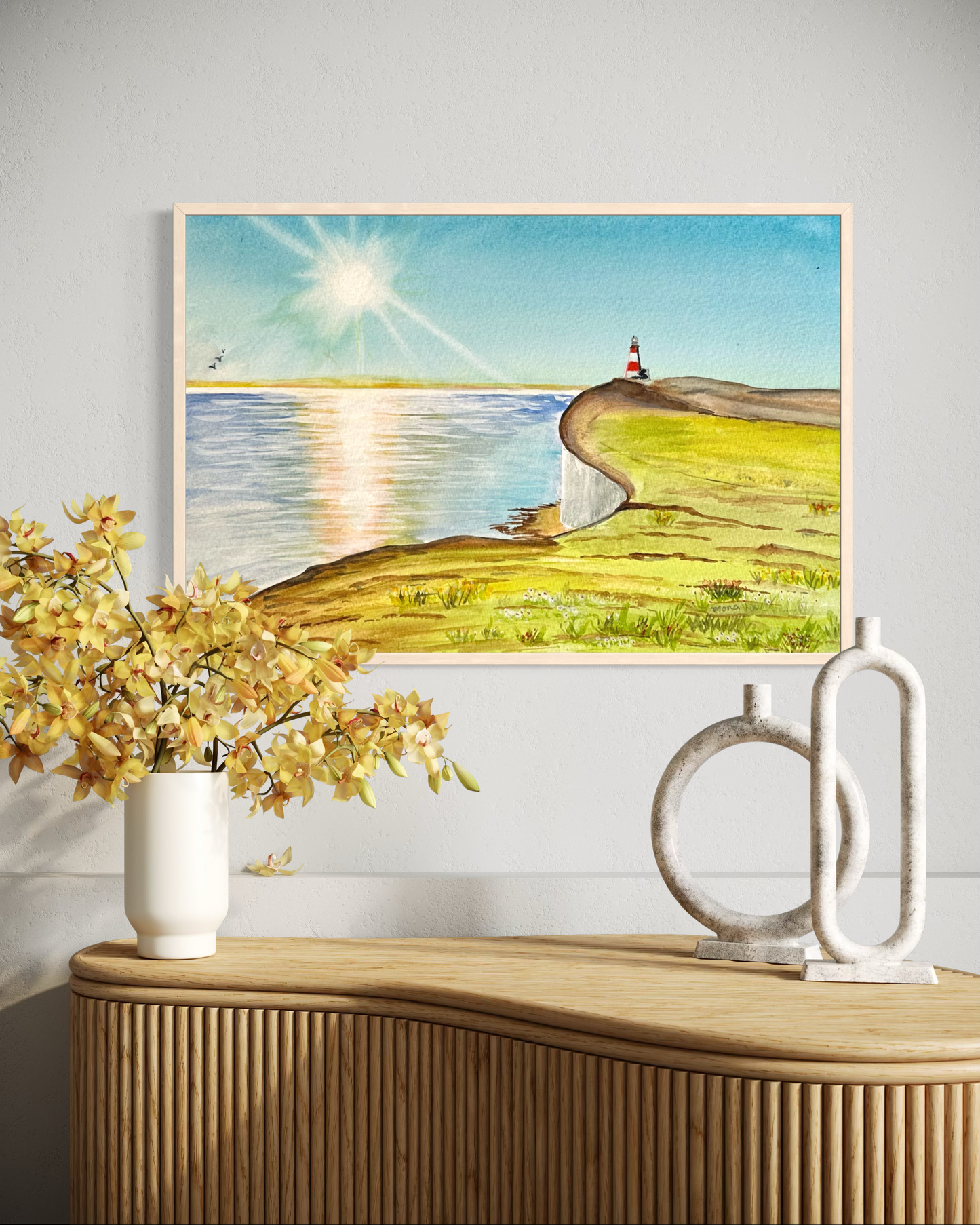 Framed artwork of a lighthouse by a lake on a wall above a wooden cabinet with decorative items.