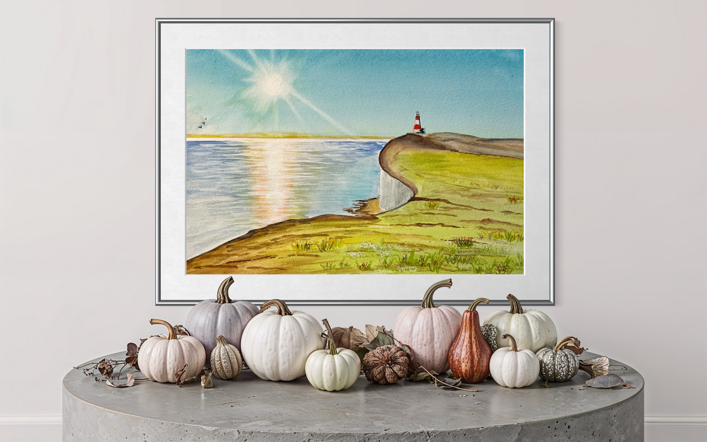 Framed artwork of a lighthouse by a lake with pumpkins on a table below.
